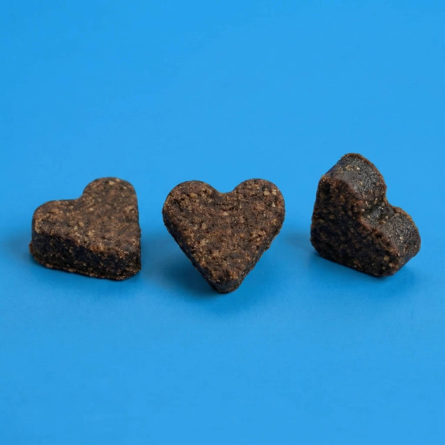 Drei dunkelbraune, herzförmige Riddys Multivitamin-Leckerlis liegen nebeneinander auf einem blauen Hintergrund. Das Bild präsentiert die Form und die natürliche Textur der Nahrungsergänzung für Katzen.