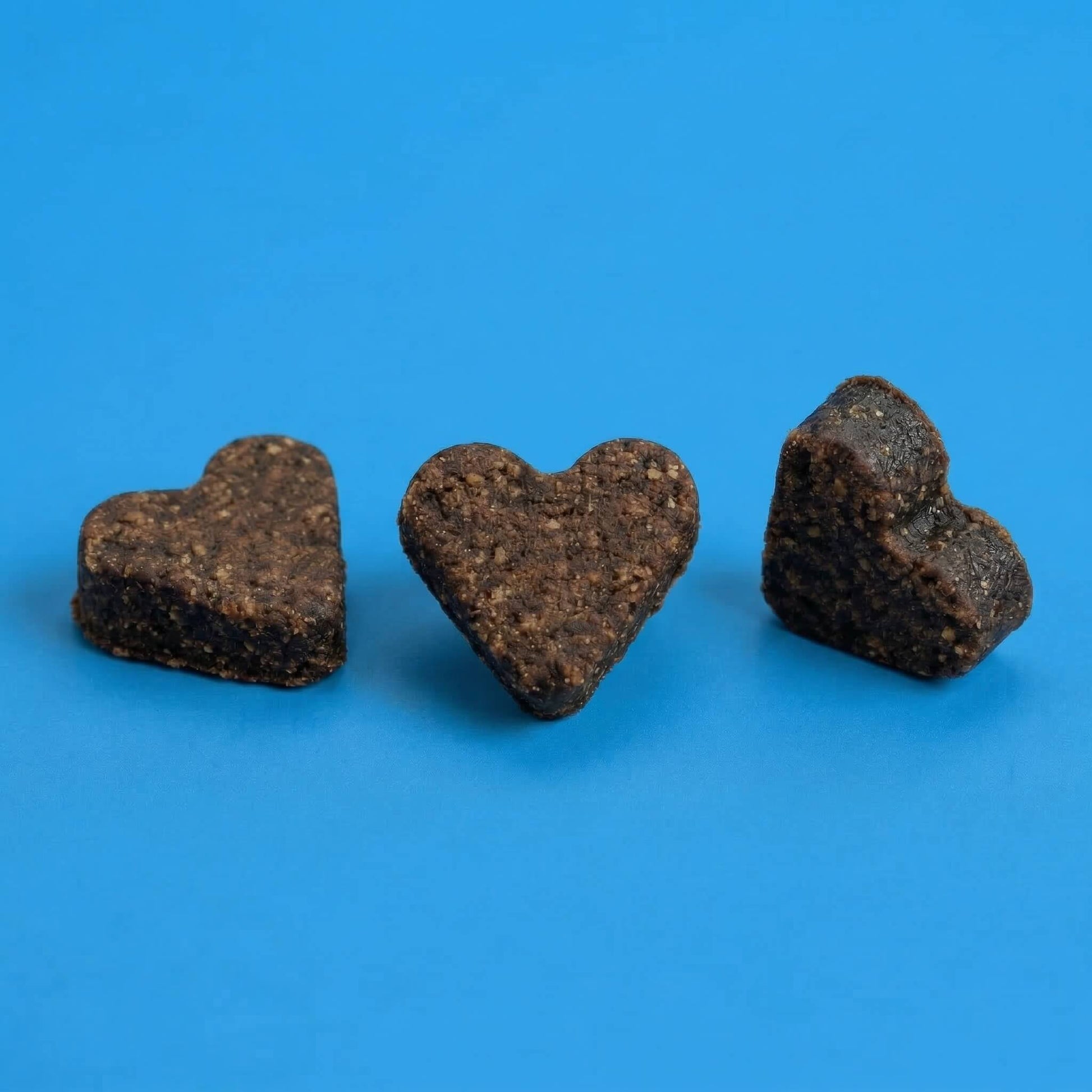 Drei dunkelbraune, herzförmige Riddys Multivitamin-Leckerlis liegen nebeneinander auf einem blauen Hintergrund. Das Bild präsentiert die Form und die natürliche Textur der Nahrungsergänzung für Katzen.