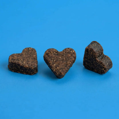 Drei dunkelbraune, herzförmige Riddys Multivitamin-Leckerlis liegen nebeneinander auf einem blauen Hintergrund. Das Bild präsentiert die Form und die natürliche Textur der Nahrungsergänzung für Katzen.
