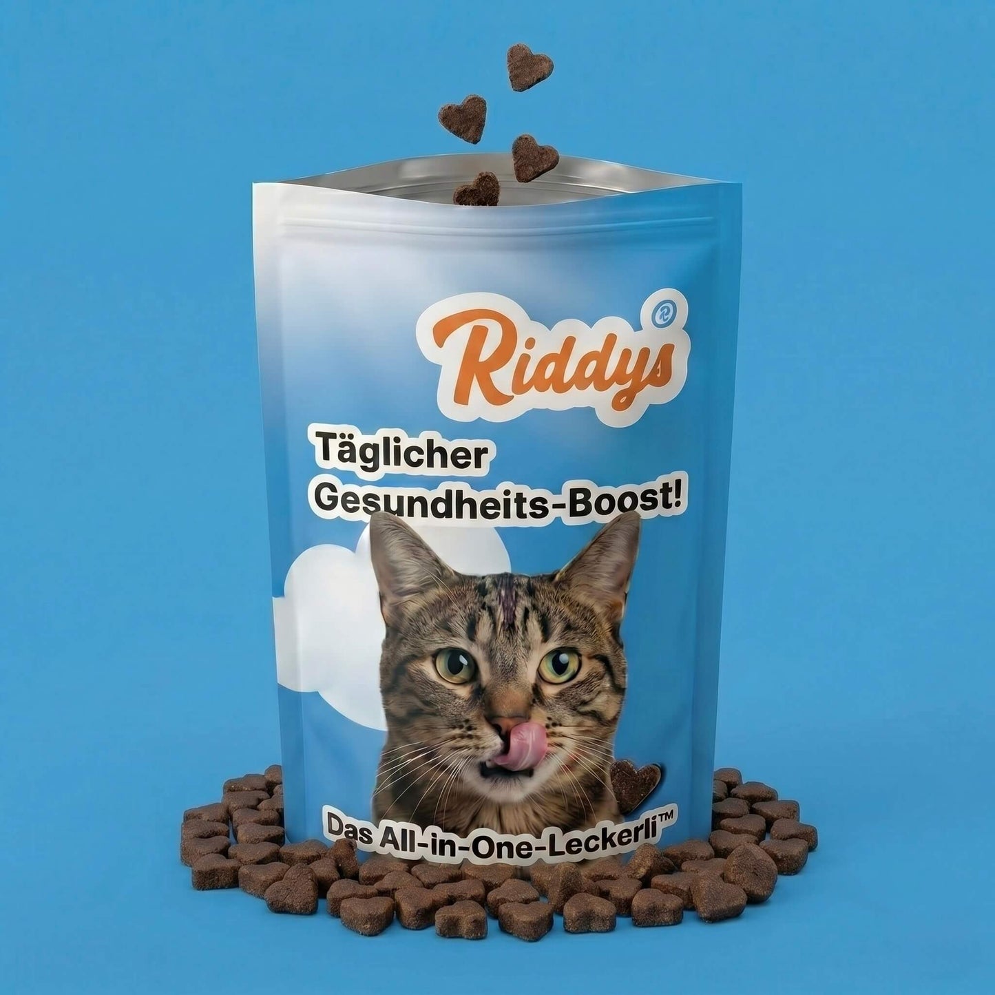 Blaue Produktverpackung der Riddys Multivitamin-Leckerlis für Katzen auf blauem Hintergrund. Die Tüte zeigt eine getigerte Katze, die sich die Lippen leckt, das Markenlogo und den Text 'Täglicher Gesundheits-Boost!'. Herzförmige, dunkle Multivitamin-Leckerlis fallen oben in die Tüte und liegen davor verstreut.